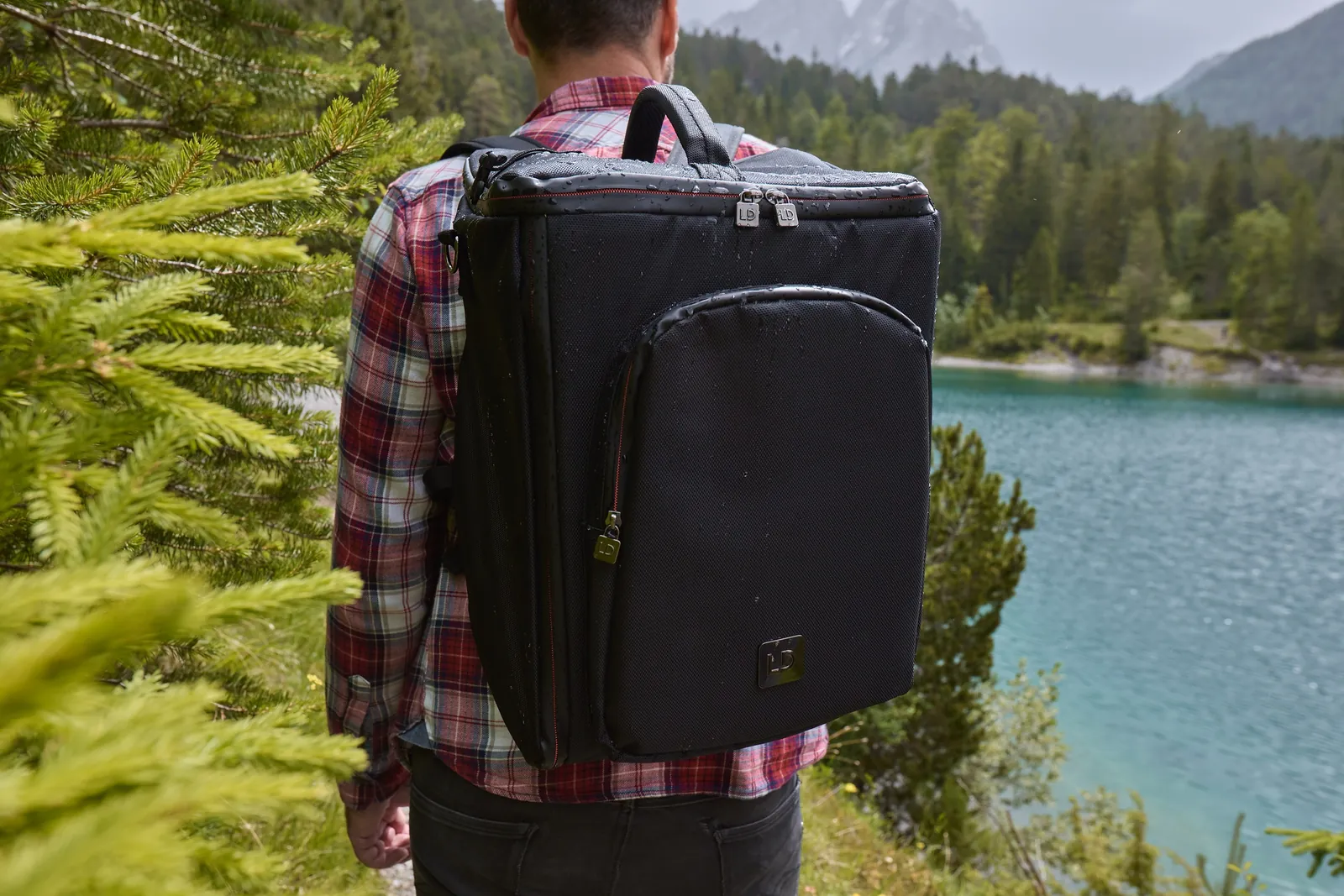 Nasser Rucksack im Close-up, kariertes Hemd, Bergsee im Hintergrund, Kampagne